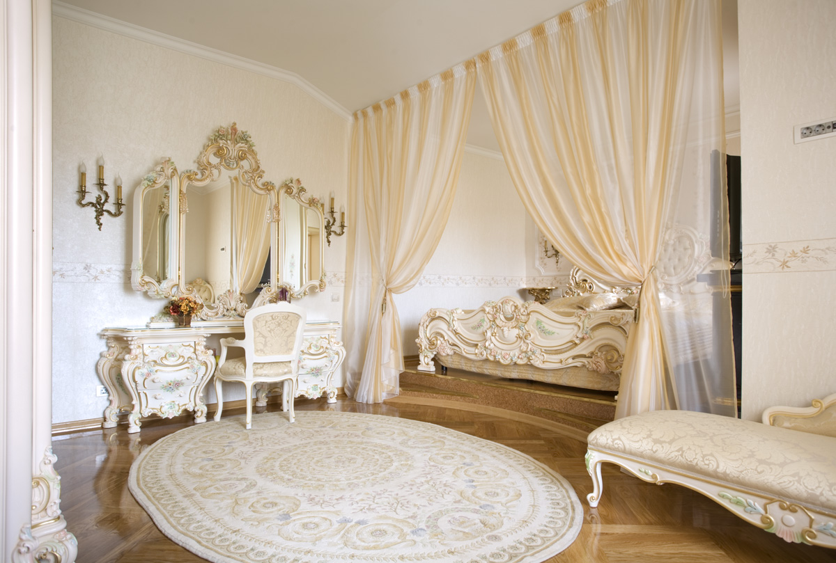 Обрамление зеркал и декоративные элементы мебели выполнены в одном стиле с использованием золота. С целью экономии пространства кровать спрятана в нишу, обрамленную шторами.